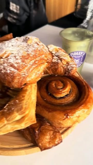 Ein Holzbrett mit Mandelcroissant, Zimtschnecke und Blätterteiggebäck, dahinter ein Iced Matcha